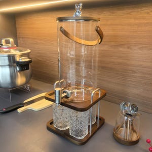 May include: A clear glass beverage dispenser with a wooden lid and a silver bear figurine on top. The dispenser has a silver spigot and sits on a wooden base with three decorative glasses. A wooden handle is attached to the dispenser.