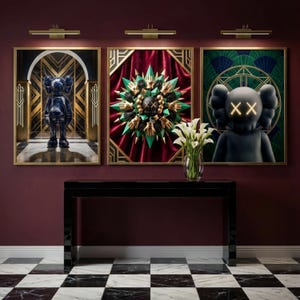 May include: Three framed art pieces with gold frames and a dark red wall. The art features a blue figure, a green and gold abstract design, and a black figure with glowing eyes. A black table and vase of white flowers are in front.