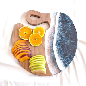 Puede incluir: Tabla de servir redonda de madera con asa. Una mitad tiene un acabado de veta de madera natural, la otra un diseño de resina azul y blanca que imita las olas del océano. Naranjas, melocotones y manzanas verdes en rodajas están dispuestas sobre la superficie de madera.