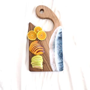 Puede incluir: Tabla de cortar de madera con asa y diseño de resina que simula olas del océano. La tabla está adornada con rodajas de naranjas, melocotones y manzanas verdes. La tabla mide aproximadamente 38 cm de largo.