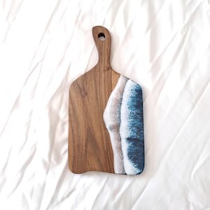 May include: Wooden chopping board with a handle. The board is half natural wood and half a blue and white ocean wave design. The board is on a white background.