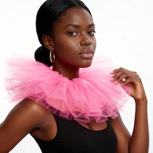May include: A woman wearing a bright pink tulle collar over a black tank top. The collar is layered and voluminous, creating a dramatic effect. The woman has dark hair pulled back and is wearing gold hoop earrings.