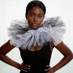 May include: A woman wearing a black tank top and gold hoop earrings, with a large, ruffled collar made of black and white tulle. The collar is layered and creates a dramatic, textured effect around the neck.