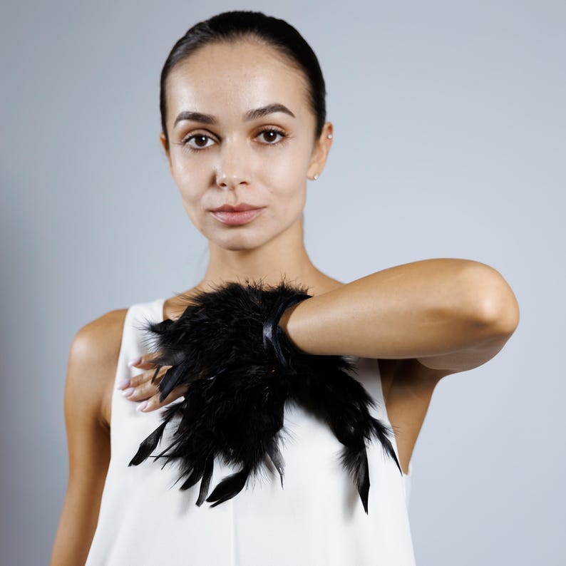 May include: A black feather cuff bracelet. The bracelet features a cluster of black feathers attached to a black band. The model is wearing a white sleeveless top.