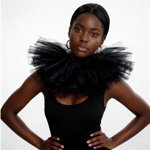 May include: A black tulle collar with multiple layers, creating a voluminous effect. The collar is worn over a black tank top. The model is wearing gold hoop earrings.