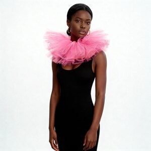 May include: A woman wearing a bright pink tulle collar over a black dress. The collar is layered and voluminous, creating a statement piece. The dress is a simple, sleeveless design. The background is plain white.