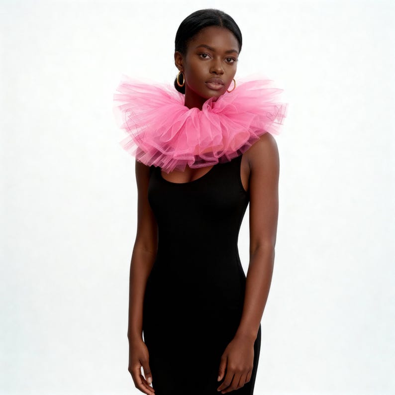 May include: A woman wearing a bright pink tulle collar over a black dress. The collar is layered and voluminous, creating a statement piece. The dress is a simple, sleeveless design. The background is plain white.