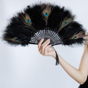 Może przedstawiać: Czarny wachlarz z piór z akcentami z piór pawia. Wachlarz ma dekoracyjny czarny uchwyt z frędzlem. Wachlarz jest otwarty, ukazując pełny kształt i wzór. Pióra pawia mają niebieskie i brązowe detale.