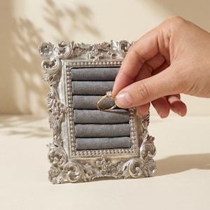 Op de afbeelding: Een zilverkleurige, sierlijke sieraden display case met een grijze fluwelen binnenkant. Een gouden ring met een kleine diamant wordt in de case geplaatst. De case heeft een decoratief, vintage-geïnspireerd frame.