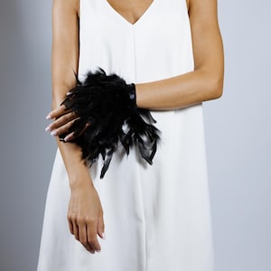 Op de afbeelding: Een zwarte veermanchetarmband. De manchet bestaat uit een zwarte band versierd met talrijke zwarte veren. De armband wordt om de pols gedragen. De achtergrond is effen lichtgrijs.