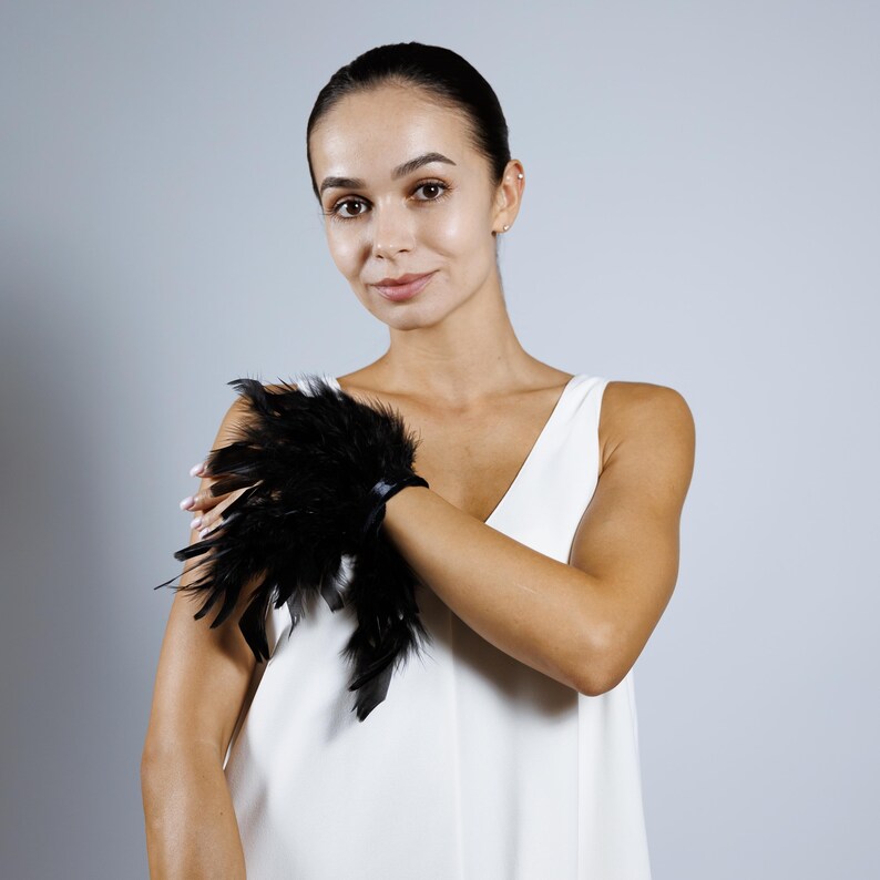 May include: A black feather cuff bracelet. The bracelet features a cluster of black feathers attached to a black band. The cuff is worn on the wrist. The background is a solid light gray.