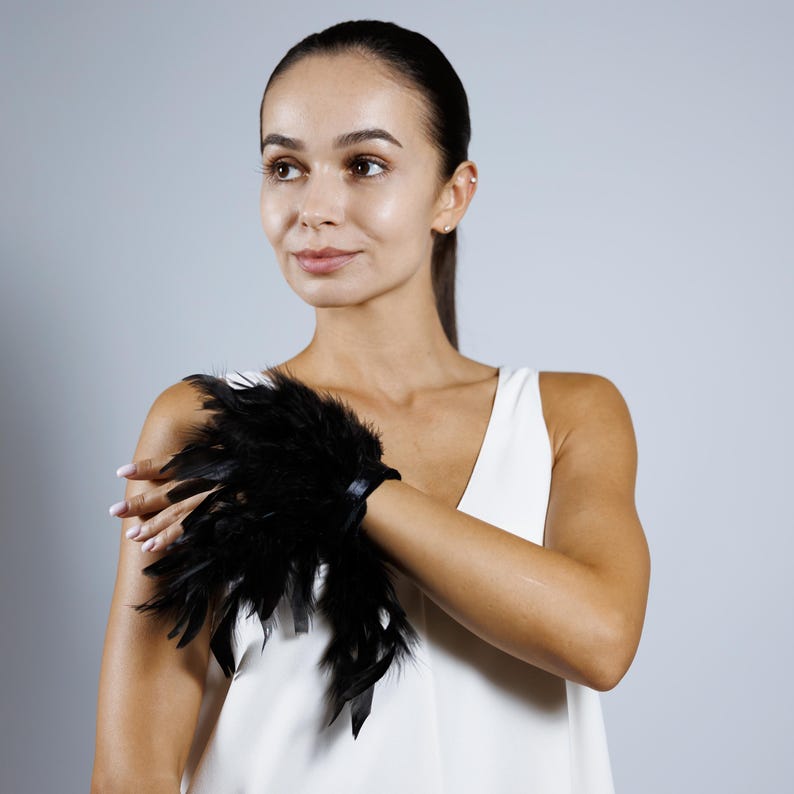 May include: A black feather cuff bracelet. The cuff is made of soft, dark feathers and is secured with a black band. The bracelet is worn on the wrist and is a striking accessory. The background is a solid light gray.