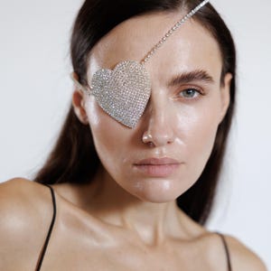 Op de afbeelding: Een hartvormig ooglapje, bezet met sprankelende kristallen, bedekt één oog. Een dunne, met kristallen bezette band loopt over het voorhoofd. Het model heeft donker haar en draagt een zwarte top met spaghettibandjes.
