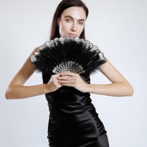 Może przedstawiać: Czarny wachlarz z piór z dekoracyjnym wzorem. Wachlarz ma czarną podstawę z czarnymi piórami i szczegółowy, ozdobny wzór pośrodku. Wachlarz jest trzymany przed czarną sukienką.