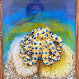 May include: A framed artwork featuring a golden octopus with blue spots, resting on a textured, off-white surface with sand and seashells. The background transitions from blue to yellow, creating a marine theme. The frame is made of wood.