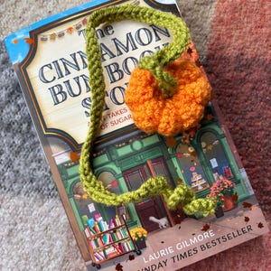 Può includere: Un segnalibro a forma di zucca all'uncinetto, arancione e verde, è appoggiato su un libro intitolato "The Cinnamon Bun Book Shop". La copertina del libro presenta un'illustrazione di una libreria. Perfetto per l'autunno.