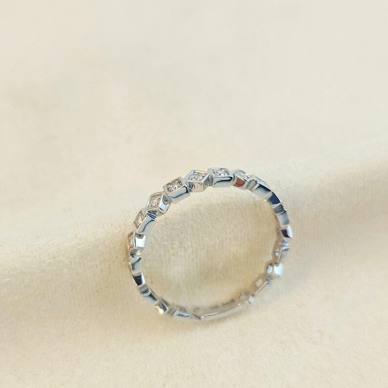 May include: A silver ring with a repeating geometric pattern. The ring is adorned with small, square-cut gemstones set within a series of interconnected shapes. The ring is shown on a neutral-toned surface, emphasising its reflective properties.