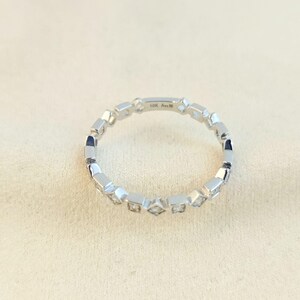 May include: A silver ring featuring a repeating pattern of square and diamond-shaped settings. The ring is set against a neutral background. The text "10K Ana M" is visible on the ring.