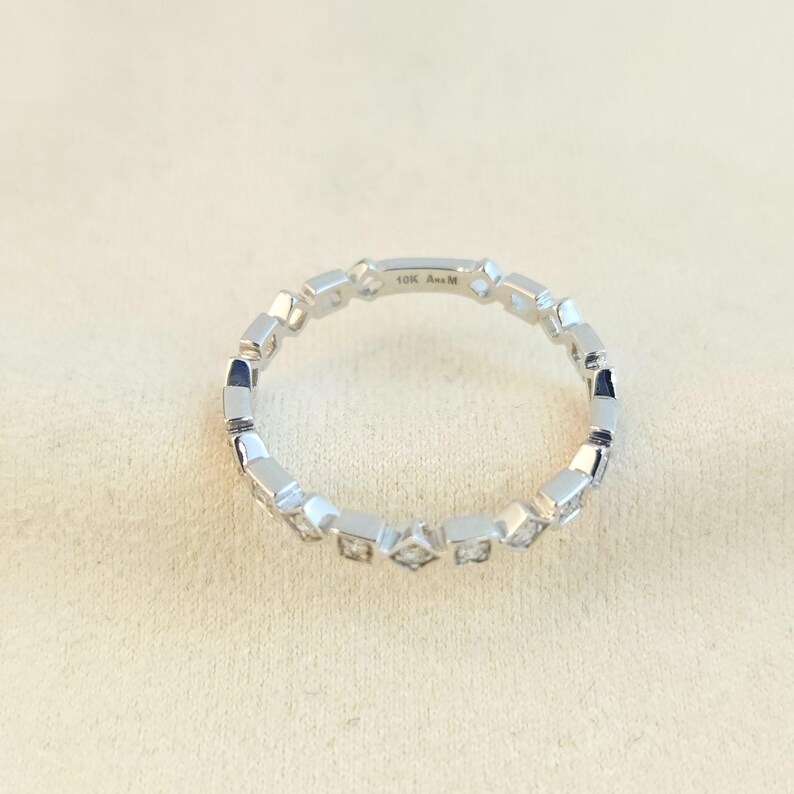 May include: A silver ring featuring a repeating pattern of square and diamond-shaped settings. The ring is set against a neutral background. The text "10K Ana M" is visible on the ring.
