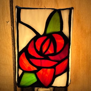 May include: A stained glass night light featuring a red rose design with green leaves and white background. The night light is rectangular with a black border and a plug at the bottom. The rose is made of various shades of red.