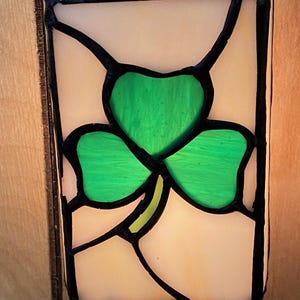 May include: Stained glass night light featuring a green shamrock design. The shamrock is set against a cream-colored background, framed by black lines. The night light is designed to plug into a wall outlet.
