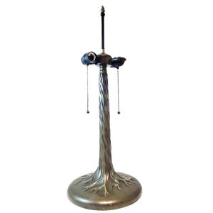 May include: A bronze-toned metal lamp base featuring a twisted, tree-trunk-like central column that flares into a wide, root-detailed circular base. The top includes two light sockets with hanging pull chains and a central finial rod for a shade.