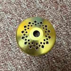 May include: A gold-colored metal object with a central hole and a decorative pattern of square and diamond-shaped cutouts. The object is circular and appears to be a decorative element or part of a larger item.