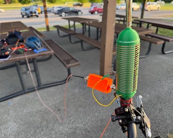 May include: A green cylindrical antenna coil with a metallic wire winding, mounted on a black handlebar assembly. An orange rectangular electronic component is connected to the coil via a green wire, with a thin red wire extending outward.