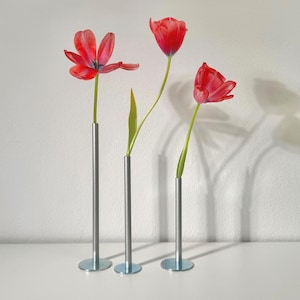 Könnte beinhalten: Drei minimalistische Metall-Blumenvasen, jede mit einer leuchtend roten Tulpe. Die Vasen haben ein schlankes, zylindrisches Design mit einem kleinen, runden Sockel. Die Tulpen befinden sich in verschiedenen Blühstadien und setzen Farbakzente.