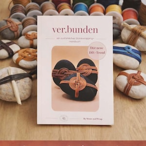 Könnte beinhalten: Ein Buch mit dem Titel "ver.bunden" und dem Untertitel "ein ausführliches Stonewrapping-Handbuch" wird gezeigt. Das Cover zeigt Steine, die mit Kupferdraht umwickelt sind. Garnrollen in verschiedenen Farben und umwickelte Steine sind um das Buch herum angeordnet.
