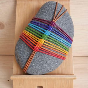 Peut inclure: Une pierre grise enveloppée de cordons aux couleurs de l'arc-en-ciel, présentée sur un support en bois clair. Les cordons sont disposés en spectre de couleurs, créant un design vibrant. Le support est en bois clair.