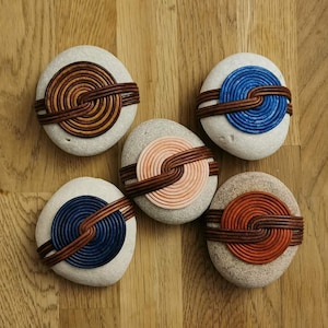 Puede incluir: Cinco piedras decorativas con diseños en espiral en varios colores, incluyendo marrón, azul, naranja y rosa. Cada piedra está envuelta con un material marrón similar al cuero. Las piedras están dispuestas sobre una superficie de madera.