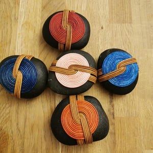 Puede incluir: Cinco piedras negras con diseños coloridos y texturizados. Cada piedra presenta una espiral central en rojo, azul, rosa o naranja, unida por bandas tejidas de color beige. Las piedras están dispuestas sobre una superficie de madera.