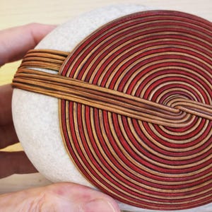 Peut inclure: Une pierre ronde et claire avec un motif en spirale en bois rouge et beige. Le bois est enroulé autour de la pierre en forme de cercle, créant un objet décoratif unique.
