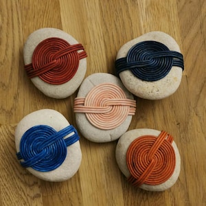 Puede incluir: Cinco piedras lisas y claras están adornadas con diseños de cordones trenzados de colores. Los cordones son en tonos de rojo, azul, naranja y rosa, creando un efecto decorativo único. Las piedras están dispuestas sobre una superficie de madera.
