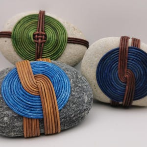 Könnte beinhalten: Drei dekorative Steine mit farbenfrohen, spiralförmigen Designs. Ein Stein hat eine grüne Spirale, ein anderer eine blaue Spirale und der dritte eine blaue Spirale. Jede Spirale ist mit braunen, lederartigen Bändern akzentuiert, was einen einzigartigen, künstlerischen Look erzeugt.
