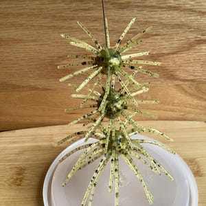 May include: A vertical fishing lure featuring three translucent, light green spherical bodies with numerous radiating, flexible spikes. Each spike and sphere is speckled with small black dots. A thin metal wire runs through the center of the spheres.