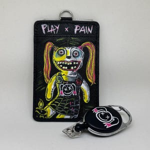 May include: Black cardholder with the text "PLAY x PAIN" and a cartoon doll design. A matching black retractable badge reel with a similar design is also shown. The doll has yellow skin, pink hair, and a black dress.