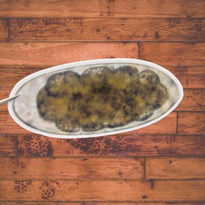 May include: An oval-shaped white ceramic serving dish containing a textured, mottled substance with shades of dark brown, olive green, and yellow. The dish has a smooth, glossy finish and a slightly raised rim, presenting a distinct contrast.