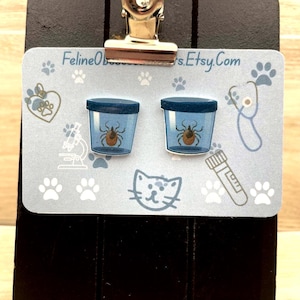 May include: A pair of stud earrings featuring a tick inside a blue container. The earrings are displayed on a light blue card with paw prints, a microscope, and the text "FelineObsessions.Etsy.Com". A silver clip holds the card.