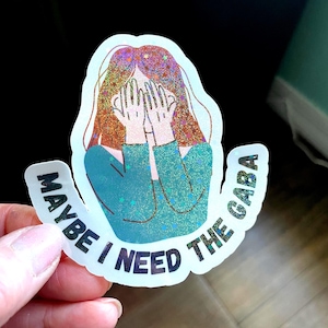 May include: A holographic sticker featuring a person with reddish-brown hair covering their face with both hands, wearing a teal long-sleeved shirt. The sticker includes the text MAYBE I NEED THE GABA in bold black letters along the bottom edge.