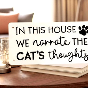 May include: A rectangular white sign with black text that reads "IN THIS HOUSE WE narrate THE CAT'S thoughts." A black paw print is in the upper right corner. The sign is on a stack of books on a wooden table.
