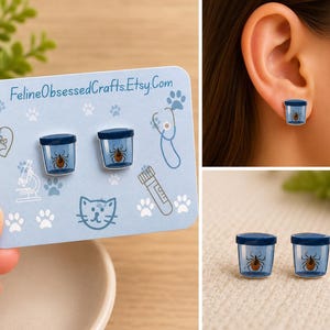 May include: A pair of stud earrings featuring a blue jar design with a tick inside. The earrings are displayed on a light blue card with paw prints, a stethoscope, and the text "FelineObsessedCrafts.Etsy.Com".