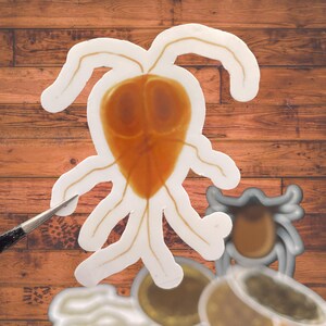 May include: A die-cut sticker featuring a brown, pear-shaped Giardia parasite with multiple flagella, positioned next to a small metal tool. Below it, a partial view of a gray tick sticker and other circular, brown-toned biological stickers.