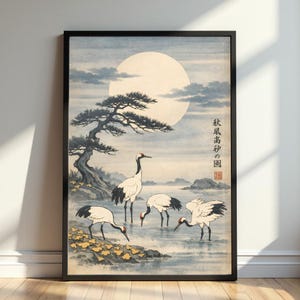 Op de afbeelding: Ingelijste kunstdruk met een Japans landschap met kraanvogels. Het kunstwerk toont vier wit-zwarte kraanvogels in het water, met een grote maan op de achtergrond. Een dennenboom en Japanse tekst zijn ook zichtbaar.
