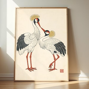 Op de afbeelding: Een ingelijste kunstdruk met twee kroonkraanvogels met witte lichamen, zwarte vleugelveren, rode gezichtsmarkeringen en gele kuiven. De vogels staan tegenover elkaar op een crèmekleurige achtergrond met een klein rood vierkant stempel.