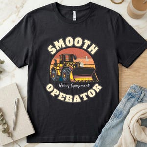 May include: A black short-sleeve t-shirt featuring a graphic of a yellow construction bulldozer centered within a sunset circle. The text "SMOOTH OPERATOR" is arched above and below the illustration, with "Heavy Equipment" written in smaller font.