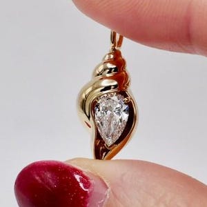 May include: A gold pendant shaped like a spiral seashell featuring a single pear-cut clear gemstone set within the centre of the shell structure. The pendant has a polished metallic finish and includes a small loop at the top for attachment.