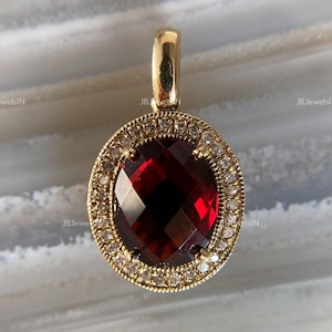 May include: An oval-shaped pendant featuring a deep red faceted garnet gemstone at the center. The stone is surrounded by a halo of small, clear pavé-set diamonds. The setting is crafted from yellow gold with a textured milgrain edge and a bail.