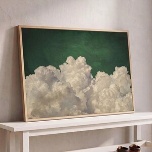 Peut inclure: Une impression d'art rectangulaire dans un cadre en bois clair représentant des nuages cumulus blancs volumineux sur un ciel vert forêt profond. L'œuvre est posée sur un banc en bois blanc avec deux bottes en cuir marron à proximité.